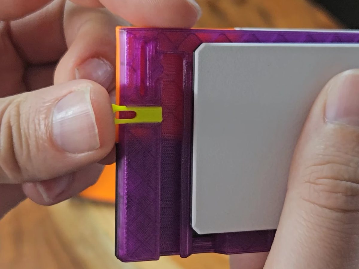 A thumb pushing a small yellow Faceplate Pin into the slot in the side edge of a translucent purple faceplate, seen from behind