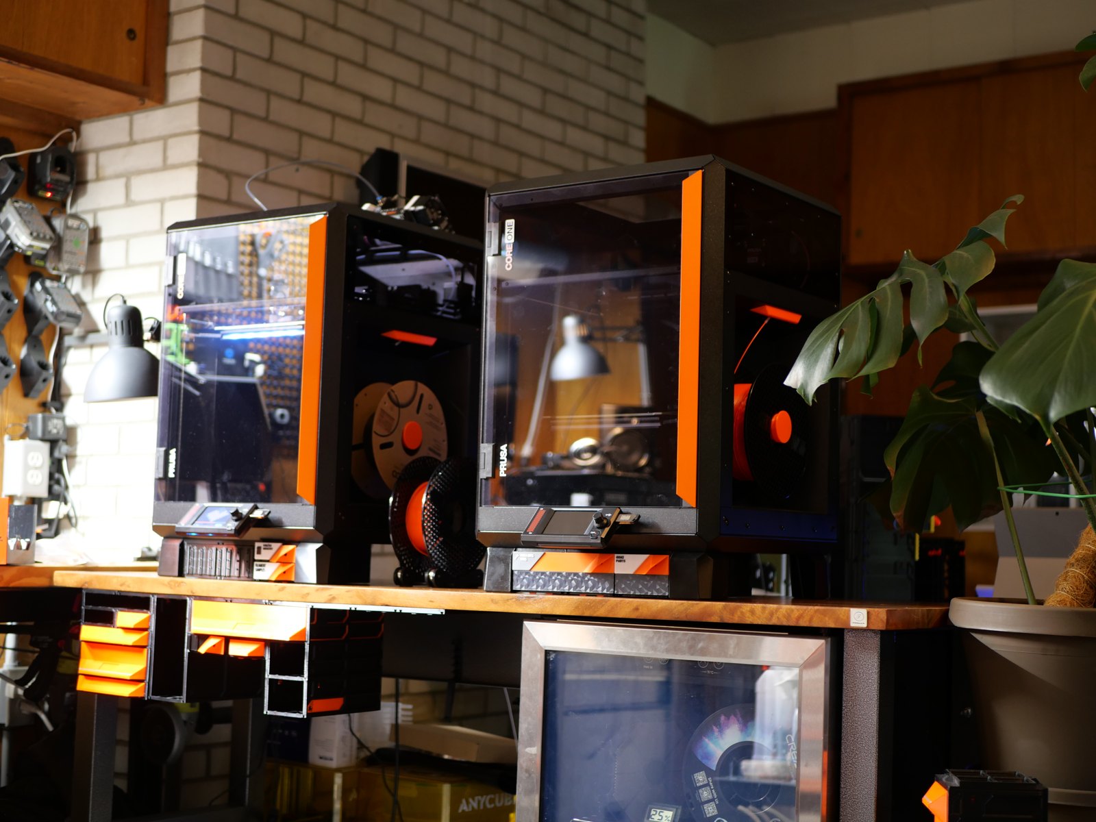 Two enclosed Prusa CORE One printers side by side on a wooden bench, with MODULITH drawers mounted under the bench top