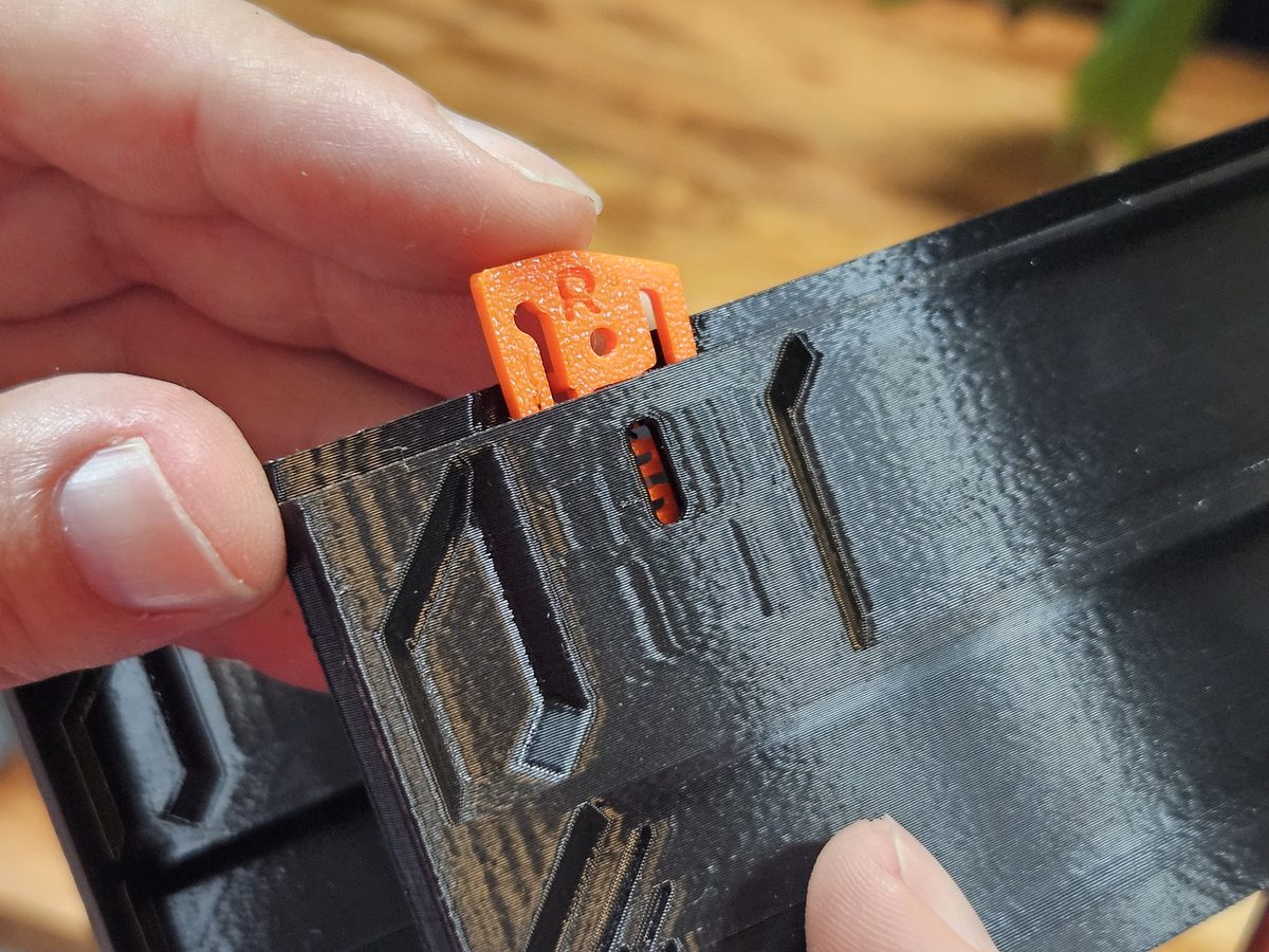 Fingers pressing an orange QuickLock marked R into the slot in the top edge of a black case wall; its spring shows through the small slot in the side of the case