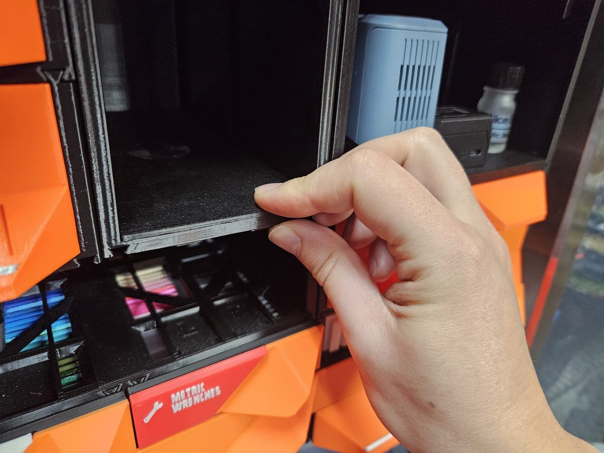 Fingers seating a black Shelf Insert into an open case, orange drawers around it