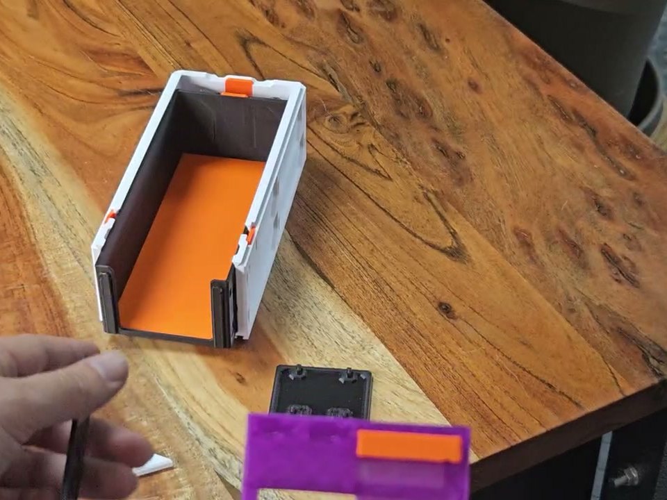 A white Decor drawer with an orange floor on a wooden bench, its front face open, a purple faceplate lying beside it