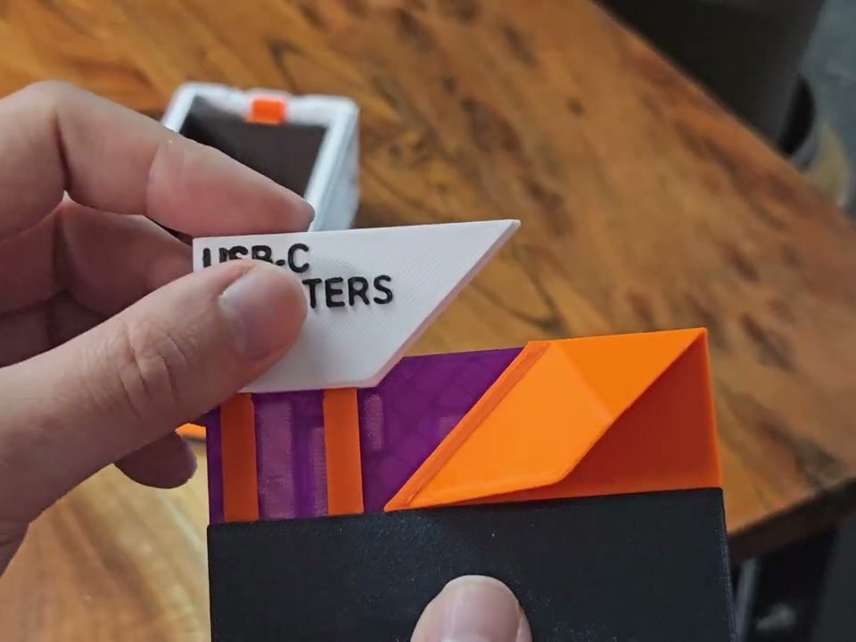 Fingers sliding a white printed label reading USB-C ADAPTERS into the angled window of a purple EdgeLabel faceplate with an orange accent