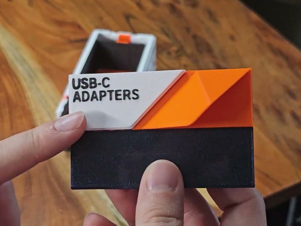 Two hands holding an assembled EdgeLabel faceplate, labeled USB-C ADAPTERS, against the open front of a white Decor drawer